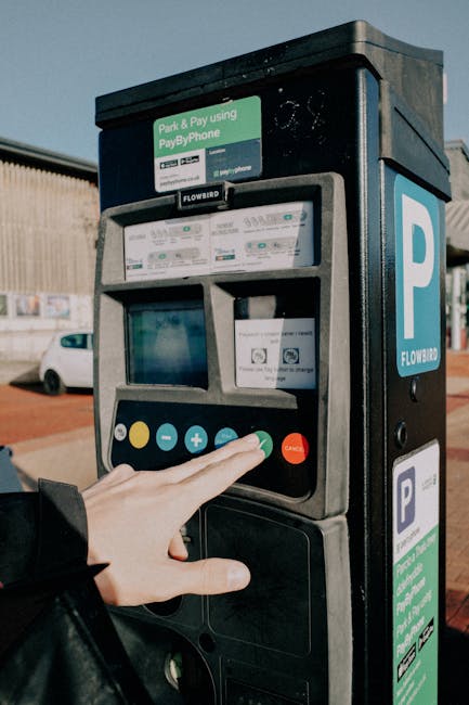 Close-up view of a person's hand pressing a green button on a parking payment machine situated in an outdoor parking area. The machine, branded Flowbird, features a small digital screen, several card 