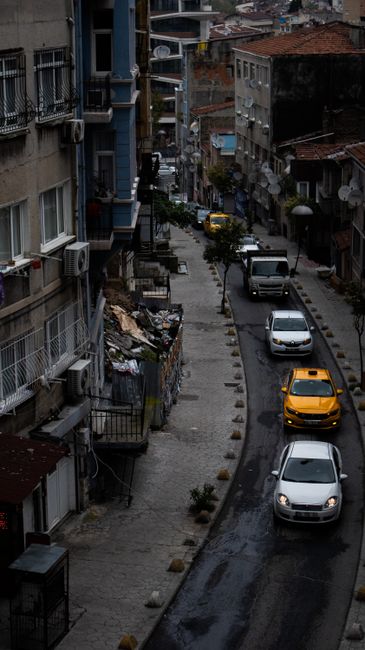 A narrow, paved street between two rows of buildings in an urban area with an overcast sky. On the left side, there are older, stone and brick buildings with small windows, while on the right side, mo