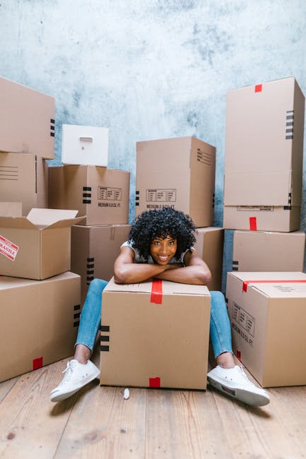 A young woman with curly black hair is sitting on the wooden floor of a room surrounded by numerous brown cardboard boxes of various sizes, some sealed with red or black tape and others partially open