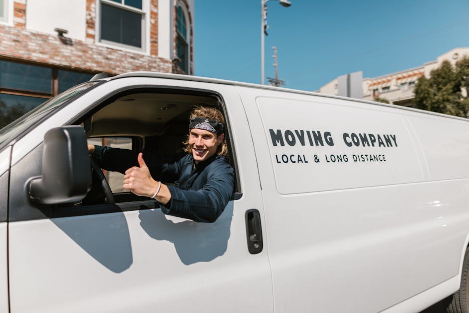 A young male moving company worker with long hair, wearing a black bandana and navy blue shirt, is sitting inside a white van labeled 'Moving Company Local & Long Distance,' giving a thumbs-up gesture