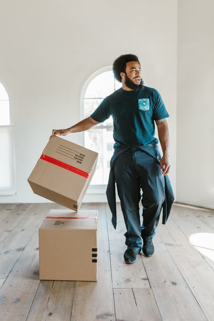 A young male moving company worker with long hair, wearing a black bandana and navy blue shirt, is sitting inside a white van labeled 'Moving Company Local & Long Distance,' giving a thumbs-up gesture