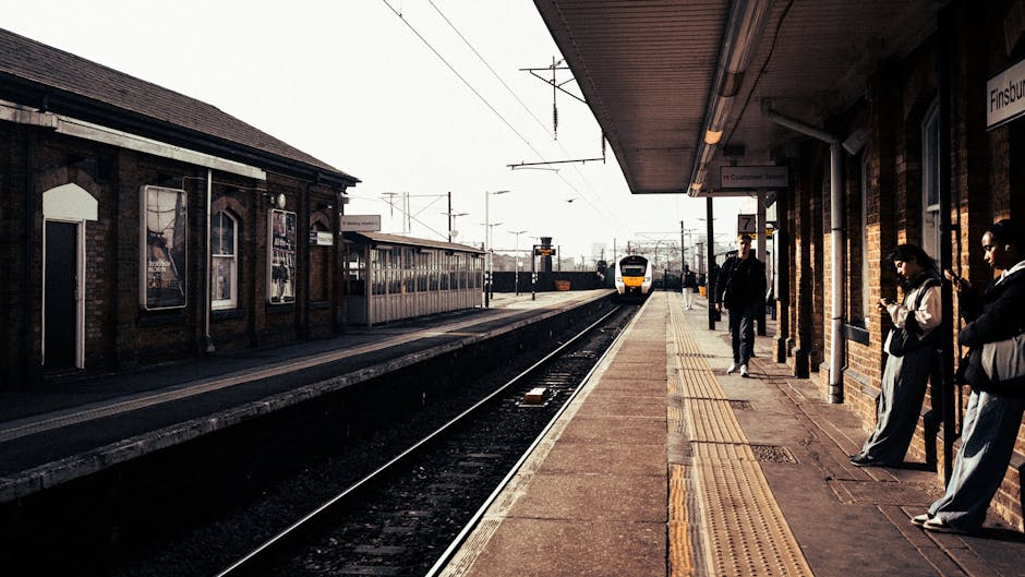 View of Finsbury Park train station platform with a group of passengers waiting near the brick wall exterior, some leaning against the wall or standing, while others are carrying or handling moving boxes and small packages wrapped in plastic and cardboard, indicating a home relocation process. A yellow and white train is approaching in the distance on the tracks, which run parallel to the platform. The platform is equipped with safety tactile paving near the edge, and a covered waiting area with a roof supported by columns. Overhead electrical wires stretch across the station, and nearby signage provides station information. The scene illustrates the loading and transit phase of furniture transport and moving logistics, with the station environment supporting smooth home relocation and packing and moving activities, as handled by Finsbury Park Removals.