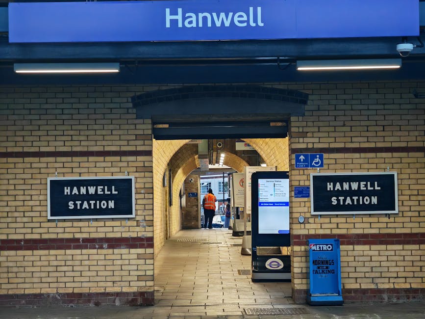 The entrance to Hanwell Station is framed by brickwork with a yellowish hue, featuring two black and white signs reading 'HANWELL STATION' on either side of a central archway. Above the arch, a blue sign with white text displays the station name. Inside, there is a tiled corridor with a person wearing an orange safety vest standing further inside, possibly involved in a home relocation or moving process. Visible on the pavement outside are a digital information kiosk, a blue Metro newspaper stand, and a blue and white accessible entrance sign indicating facilities for disabled travelers. The scene suggests a setting where furniture transport and packing activities might occur, with station access tips relevant for residents planning removals or home moves.