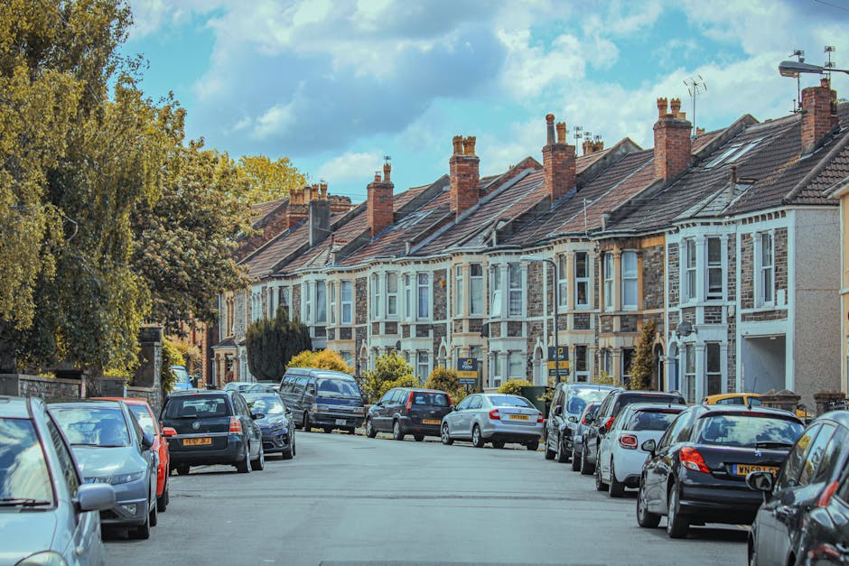 A residential street with a row of terraced houses featuring brick facades and bay windows, lined with parked cars along the pavement. The street is partly shaded by large trees with green foliage, and the sky above is partly cloudy with patches of blue. Visible in the foreground are various vehicles, including sedans, hatchbacks, and a van, parked both on the street and on the pavement. The scene captures a typical home relocation area where Finsbury Park Removals manages logistics such as parking and access for furniture transport, with the pavement dispersed with cardboard boxes, packing blankets, and other moving materials for packing and loading goods into vans. The overall environment suggests an organized moving process underway, with elements indicating the preparation for house removals, including loading and transportation actions relevant to residential moves.