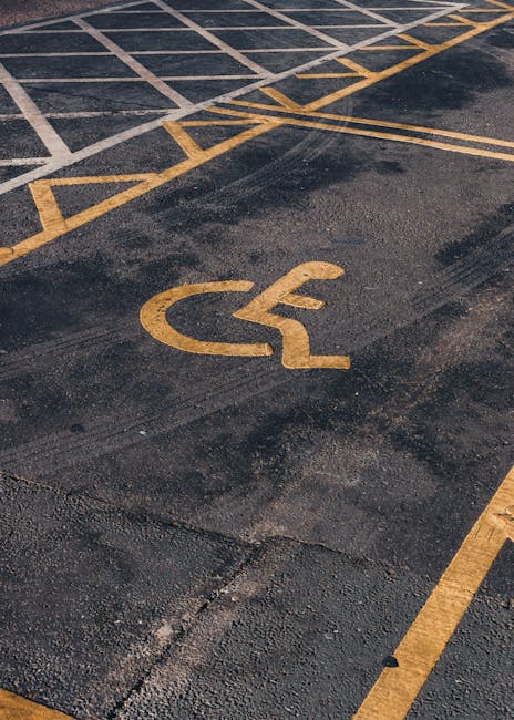 A close-up view of an outdoor parking area featuring a yellow wheelchair symbol painted on the dark asphalt surface, indicating accessible parking. Surrounding the symbol are yellow box junction markings and parking lines, with some of the lines slightly worn. The scene is illuminated by natural light, and the area appears to be adjacent to a residential or commercial building, suggesting a location suitable for home relocations or furniture transport. The image reflects typical parking arrangements used during house removals to facilitate easy access for moving vehicles and aid in the loading and unloading of furniture, boxes, and packing materials. Finsbury Park Removals* may conduct operations in such parking zones when assisting with house removals and moving services on Stroud Green Road, ensuring coordinated access for efficient relocation logistics.